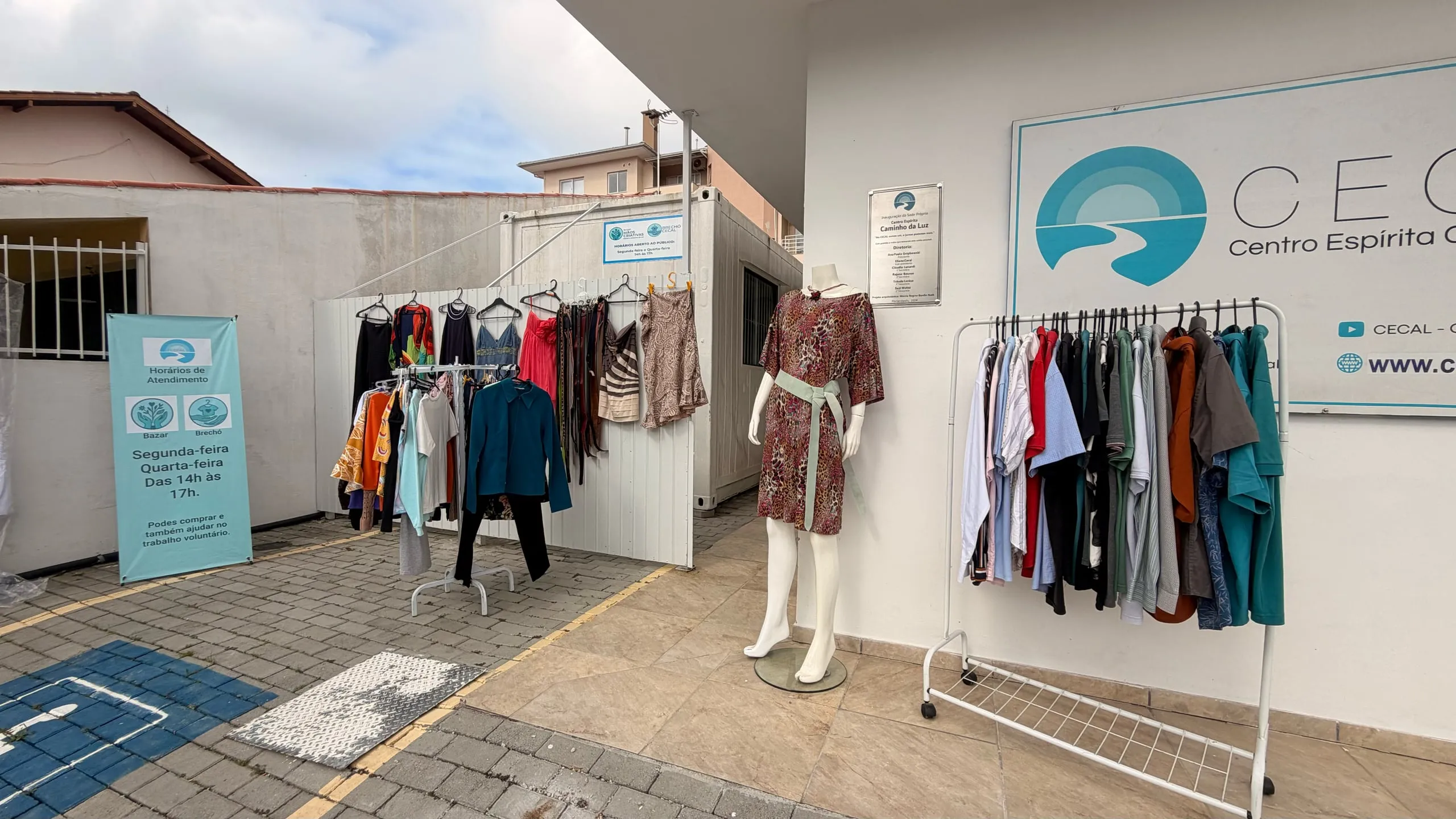 O brechó montado na entrada da casa: araras com roupas, um manequim de vestido e, ao fundo, a placa do CECAL e o banner com os horários.