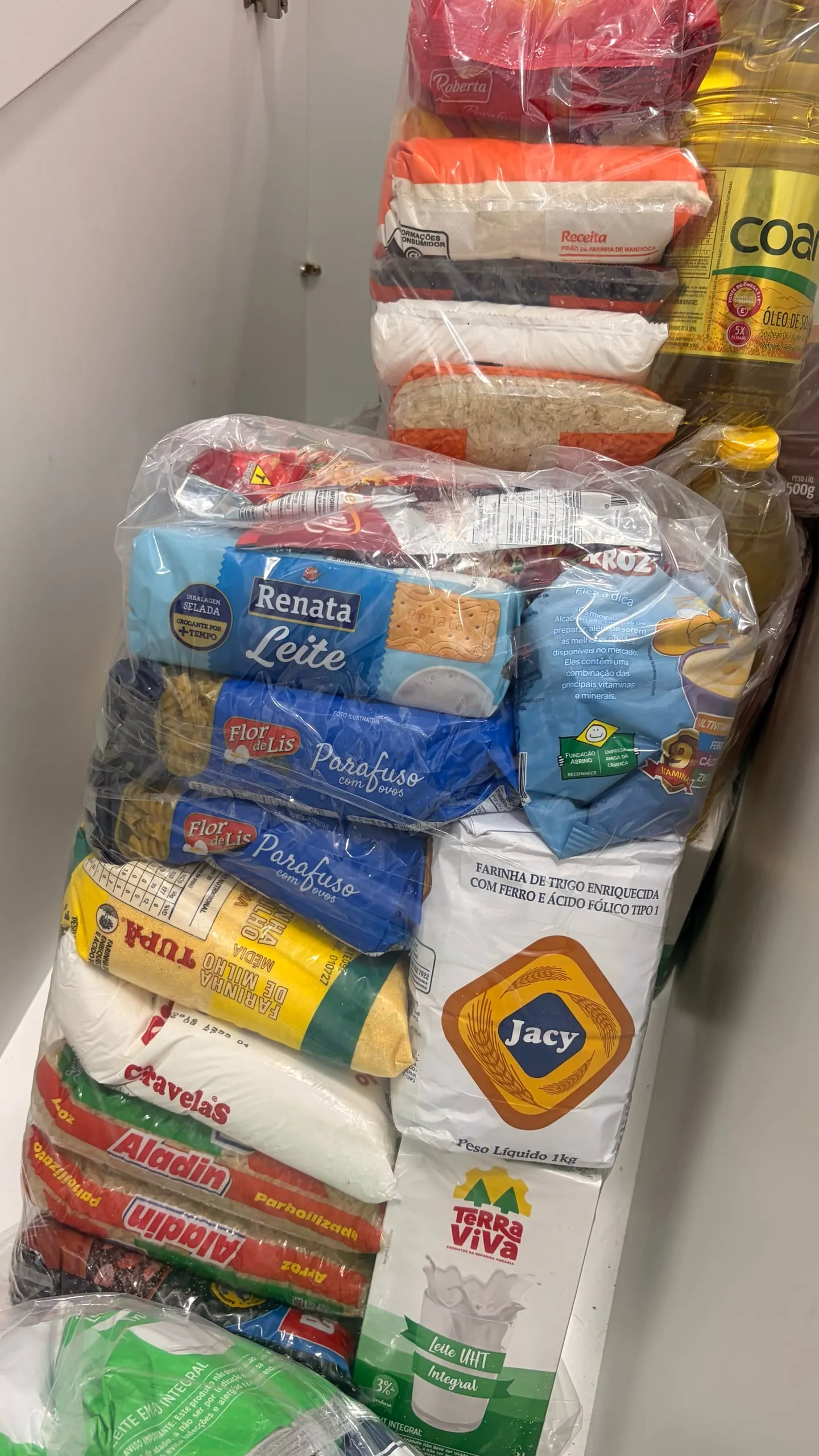 Doações de alimentos empilhadas num armário da casa: pacotes de arroz, feijão, macarrão, farinha, leite e óleo.