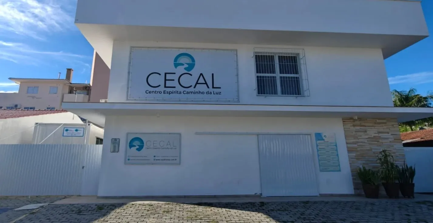Fachada da sede do CECAL vista de frente: prédio branco de dois andares com a placa do Centro Espírita Caminho da Luz, o portão e o céu azul.
